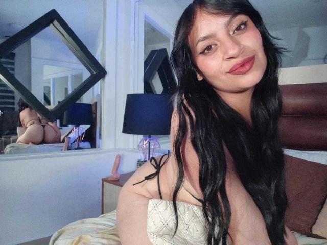 AlexaMiranda with long black hair, smiling, wearing a white thong, on a bed, reflecting in a diamond-shaped mirror, showing her bare ass in the reflection.