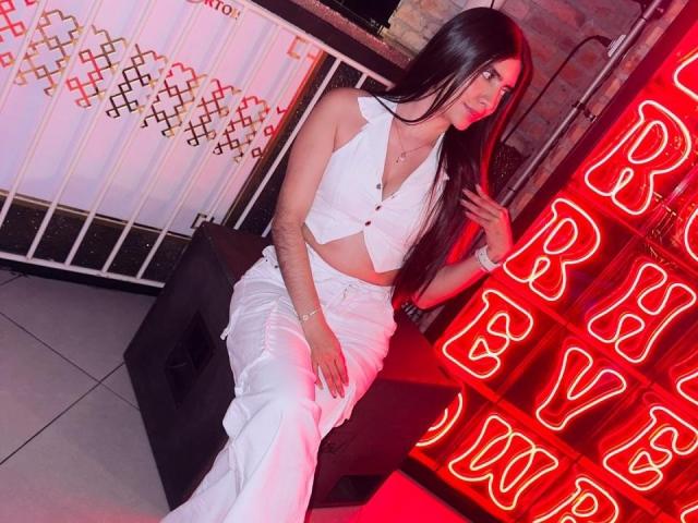 LuanaJoys with long black hair, wearing a white crop top and pants, sitting on a black stool, neon "B" signs in red, white wall with geometric patterns.