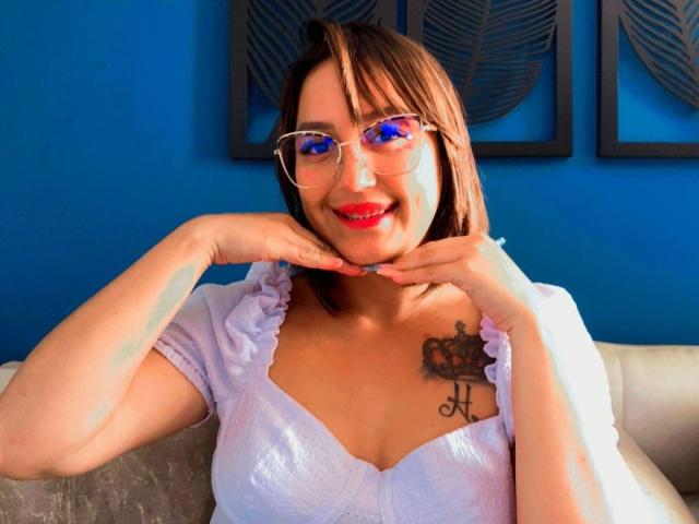 AnneLucia with red lipstick, glasses, and a white ruffled top, showing off her tits and a crown tattoo on her right boob, smiling with hands on her cheeks.
