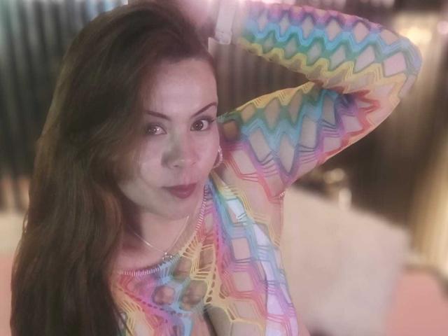 AstridBayon with long brown hair, wearing a colorful, zigzag-patterned, see-through top, raising her right arm, smiling slightly, pink lipstick, and a silver necklace.