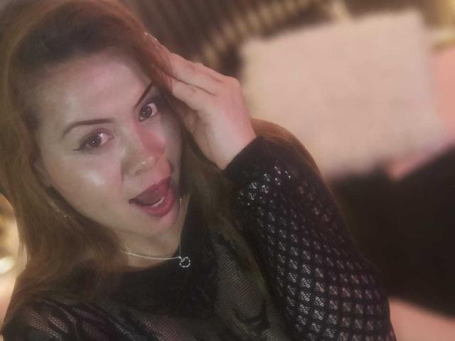 AstridBayon, com pele brilhante, cabelo castanho e blusa de malha preta, com a mão no cabelo, boca aberta, olhando para a câmera, usando um colar pequeno, fundo desfocado..