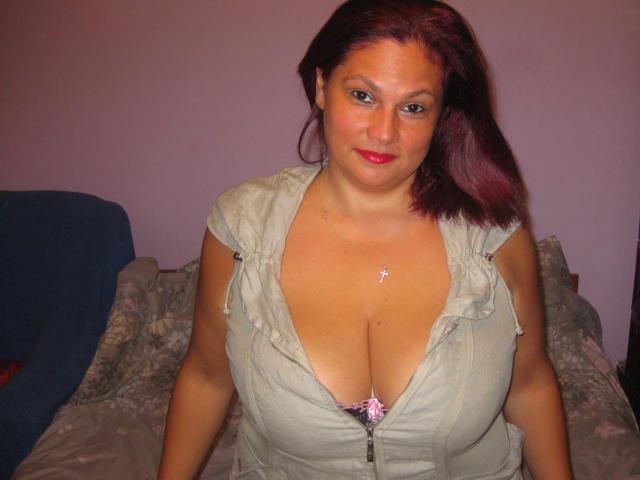 HellenBooty69 hat große Brüste, roten Lippenstift und dunkelrotes, mittellanges Haar, trägt ein graues Kleid mit tiefem Ausschnitt, auf dem ein rosafarbenes Schleifchen zu sehen ist, und eine Kreuzkette..