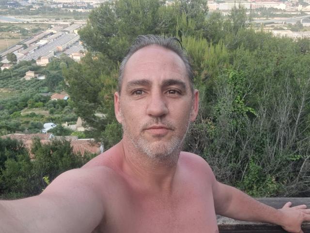Shirtless ManiSexy69 with gray beard, short brown hair, and slight tan, holding railing, green bushes in background, serious expression, arms resting on edge.