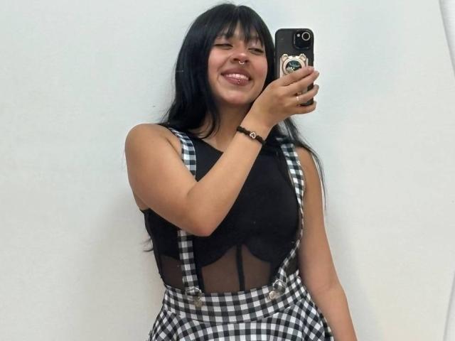 Mia Whitmore, con cabello negro, sonriendo, vistiendo una blusa de malla negra, tirantes a cuadros negros y blancos, sosteniendo un teléfono, mostrando senos pequeños, y llevando una pulsera negra..