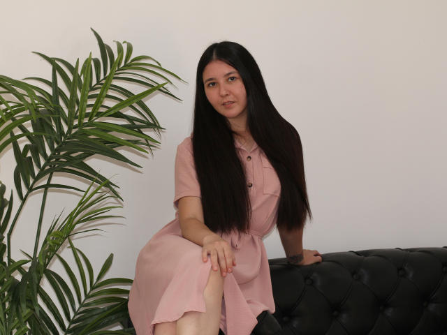 Liza Holmes, con i suoi lunghi capelli neri, il vestito rosa e il corpetto abbottonato, è seduta su un divano nero imbottito, appoggiata alla gamba sinistra, con una pianta verde alla sua sinistra. Ha un'espressione carina e sicura di sé..