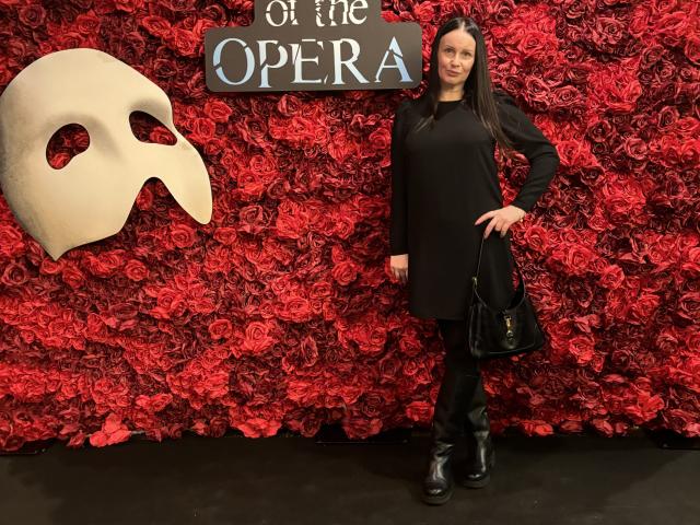 JasmineVelvet stands in front of a red rose wall, wearing a black dress, black boots, and a black handbag, with a white mask to the left.