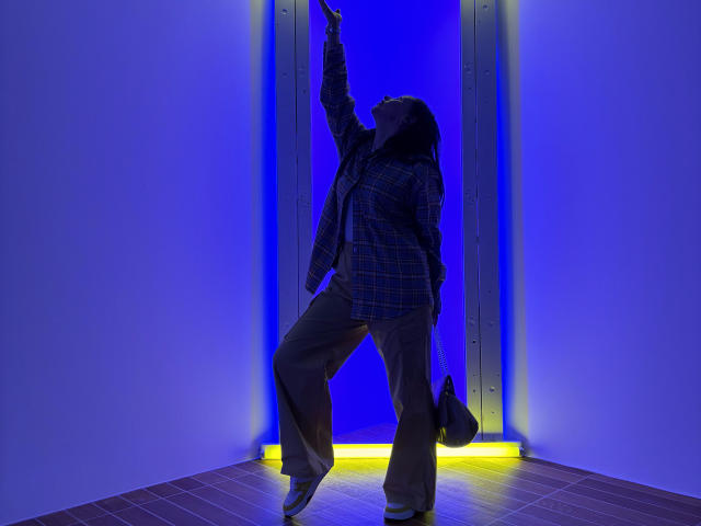 JasmineVelvet with dark hair, plaid jacket, and loose pants, pointing upward, standing on one leg, in a blue-lit doorway, holding a black bag, looking up.