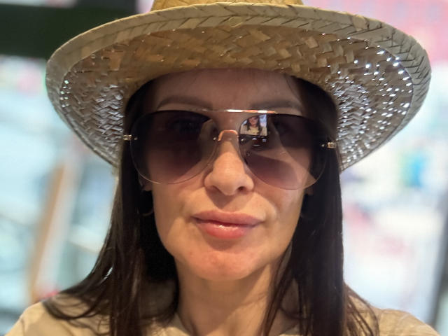 JasmineVelvet wearing a straw hat, big brown sunglasses, and a grey shirt, with long dark hair and a slight smile, looking straight at the camera.
