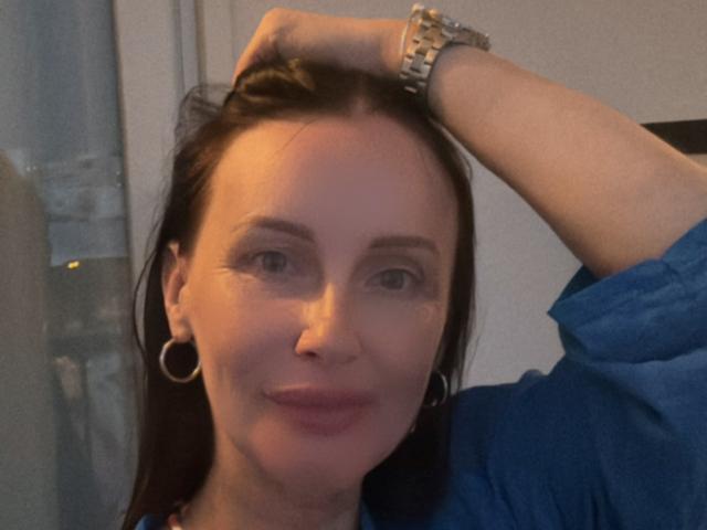 JasmineVelvet with dark brown hair, blue eyes, wearing a blue shirt, hoop earrings, and a silver watch, hand in hair, slightly smiling, soft light, casual selfie.
