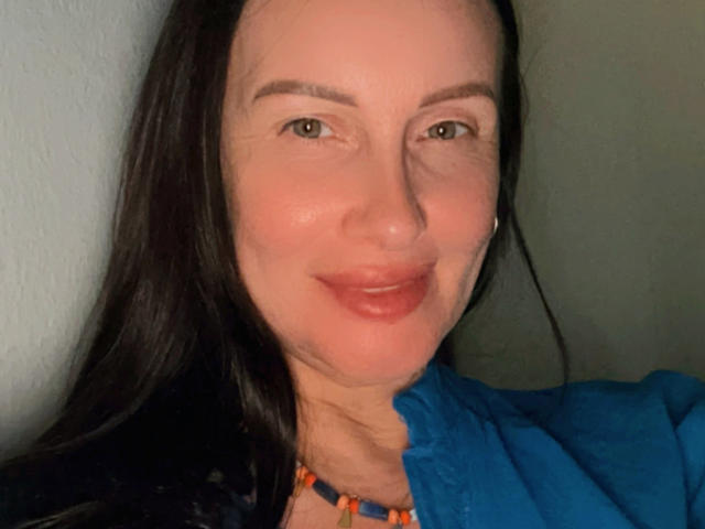 JasmineVelvet with long black hair, light skin, subtle smile, brown eyes, wearing a blue shirt and colorful bead necklace, looking at camera with soft lighting.