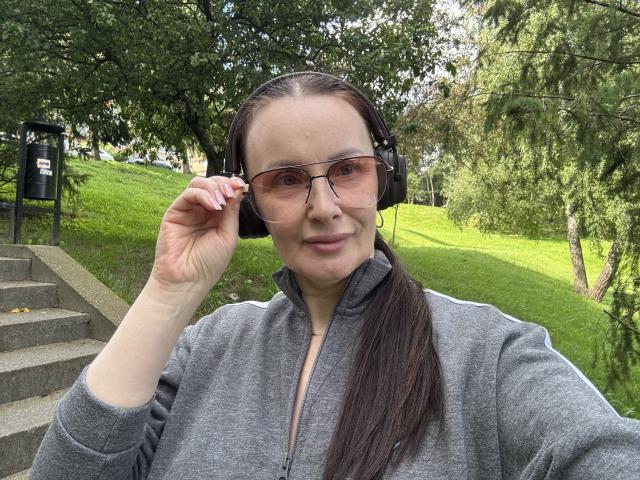 JasmineVelvet with her ass-kicking headphones on, wearing glasses and a gray zip-up, standing in a green park with a concrete staircase, adjusting her headphones with a smile.