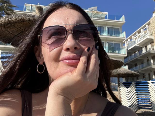 JasmineVelvet with long dark hair, pink-tinted aviator sunglasses, hoop earrings, black nail polish, brown tank top, hand on cheek, blue sky, white beach buildings in background.
