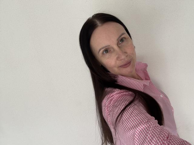 JasmineVelvet has long dark brown hair, light skin, green eyes, and wears a red checkered shirt, smiling slightly with her head tilted to the right.