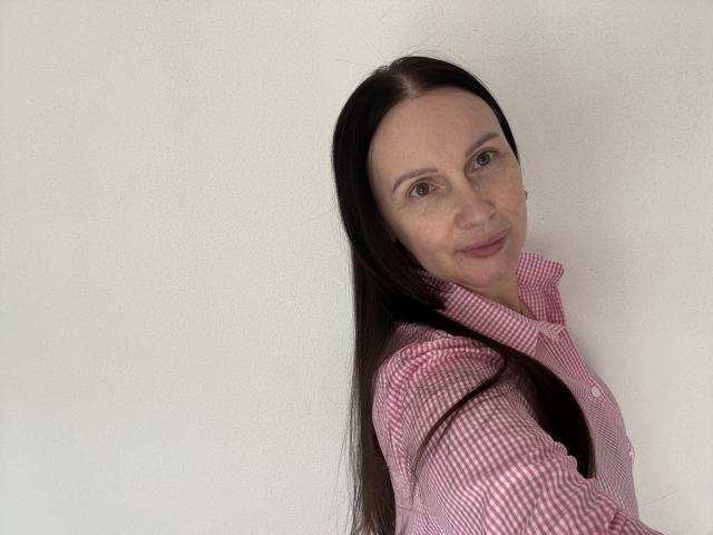 JasmineVelvet with long brown hair, wearing a pink checkered shirt, looking over her shoulder with a slight smile, showing her small tits against a white wall.