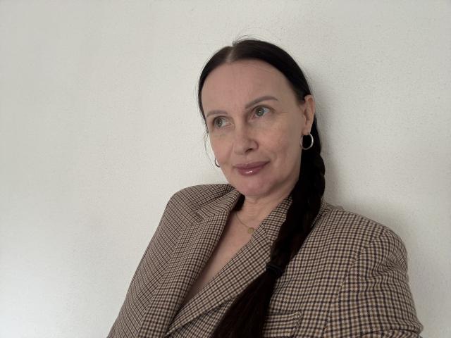 JasmineVelvet has pale skin, green eyes, and a slight smile. She wears a brown checkered blazer and has a long black braid, with hoop earrings.