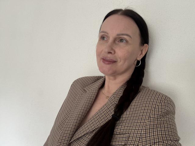 JasmineVelvet with brown hair in a braid, wearing a checkered blazer, small hoop earrings, and a subtle smile, leaning against a white wall.