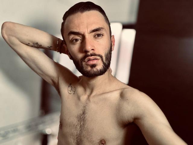Jake Magnus senza camicia, con una barba curata, capelli scuri e pettinati all'indietro, un tatuaggio sul braccio destro e un piccolo tatuaggio con il simbolo della pace sul petto sinistro, con una mano dietro la testa, che guarda verso la fotocamera..
