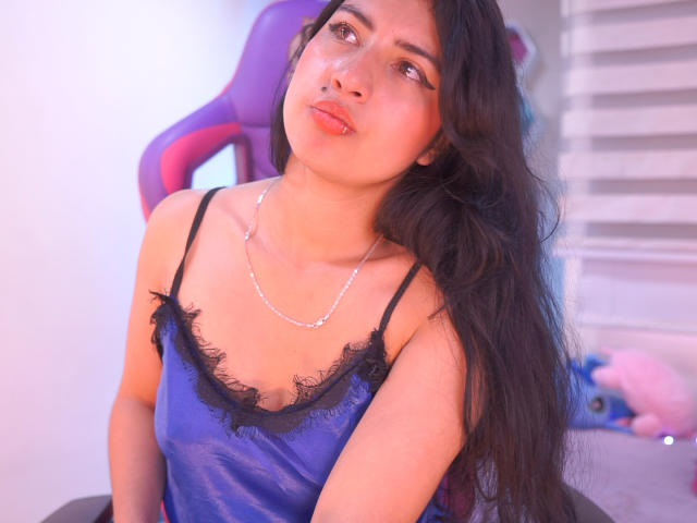AlexaGrey69 with long black hair, pouty pink lips, blue satin camisole with black lace, small tits, silver necklace, looking up, sitting on purple chair, soft pink light.