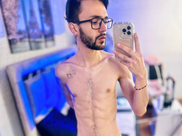 JakeMagnus, shirtless, with a slim, hairy-chested build, holding a phone, wearing glasses, has a tattoo on his right shoulder, and a beard, taking a mirror selfie.