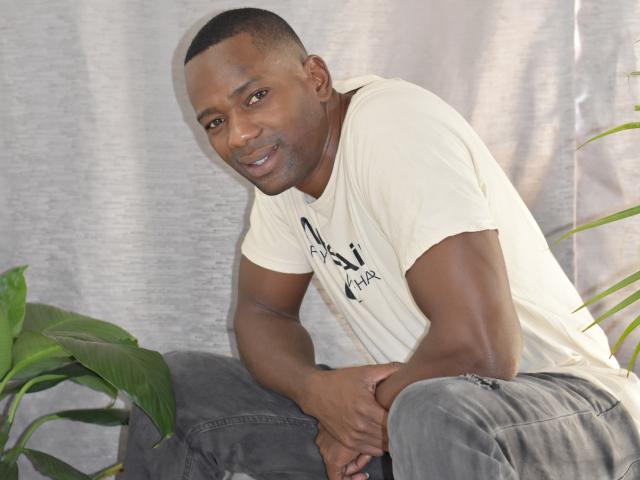 AntonyCruze, dark-skinned dude with short buzzcut, wearing cream tee and gray pants, leaning forward, hands on knees, green plant on left, smiling, looking at camera.