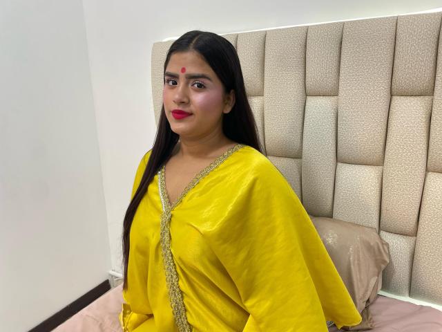 NayaYalitza has dark brown hair, red lipstick, and a yellow traditional dress with gold trim. She's sitting on a bed with a beige tufted headboard.