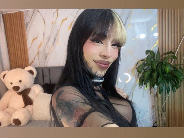 AnneKisss with black hair, tattoos, and a choker, smiling, showing her tits through a sheer top, next to a white teddy bear and a green plant.