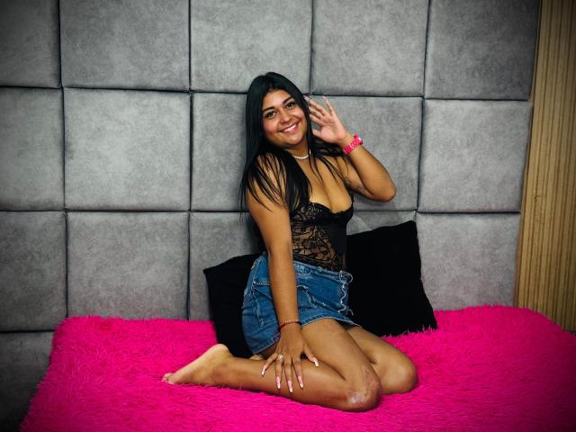 SilvanaNoir with long black hair, smiling, wearing a black lace top, showing her small tits, and blue denim shorts, kneeling on a pink fuzzy bed, touching her ear.