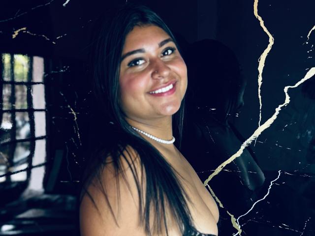SilvanaNoir with long black hair, smiling, wearing a pearl necklace, and showing her bare tits, with a dark, crackled background. She has a cute, sexy face.