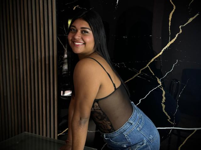 SilvanaNoir with long black hair, smiling, wearing a sheer black top and blue jeans, leaning forward, showing her ass and black lace pattern on her back.