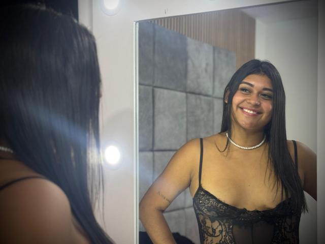 SilvanaNoir with long black hair, smiling at herself in a mirror, wearing a black lace lingerie top, showing small tits, and a pearl necklace, in a well-lit room.