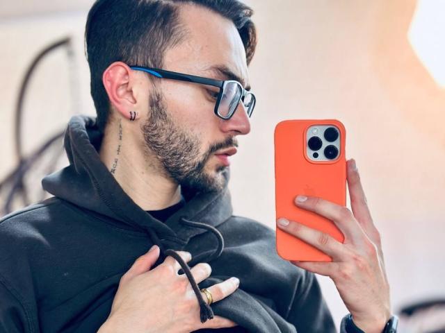 JakeMagnus with glasses, short brown hair, beard, black hoodie, holding orange phone, looking at screen, ring on right hand, tattoo on left neck, focused expression.
