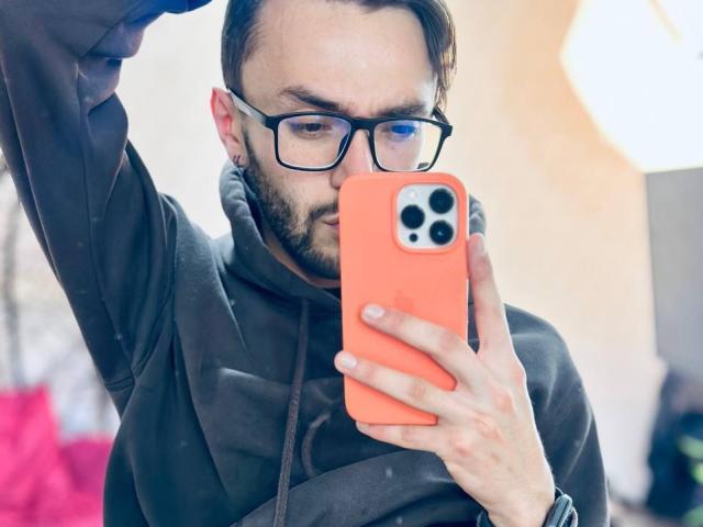 JakeMagnus with glasses, beard, and brown hair, holding an orange iPhone, taking a selfie in a mirror, wearing a dark hoodie, right arm raised.