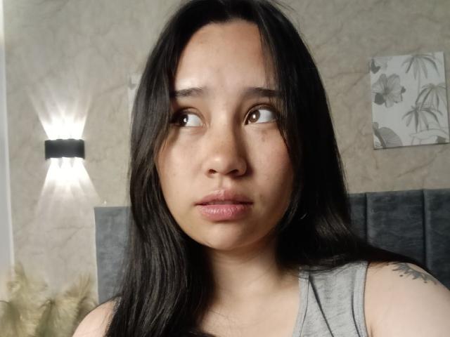 Karollteyes has medium brown eyes, long black hair, and a neutral expression. She's wearing a gray tank top, with a small tattoo on her right shoulder.