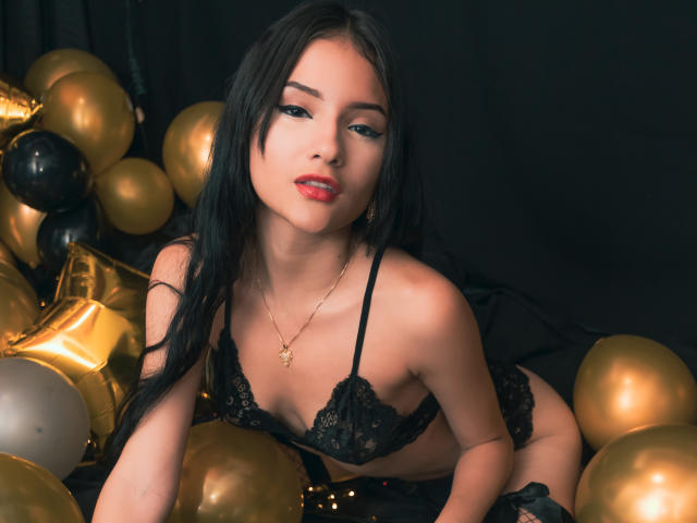 LizzeJoy with black lace bra and panties, gold necklace, red lips, and long black hair, surrounded by gold and black balloons, leaning forward with a seductive look.