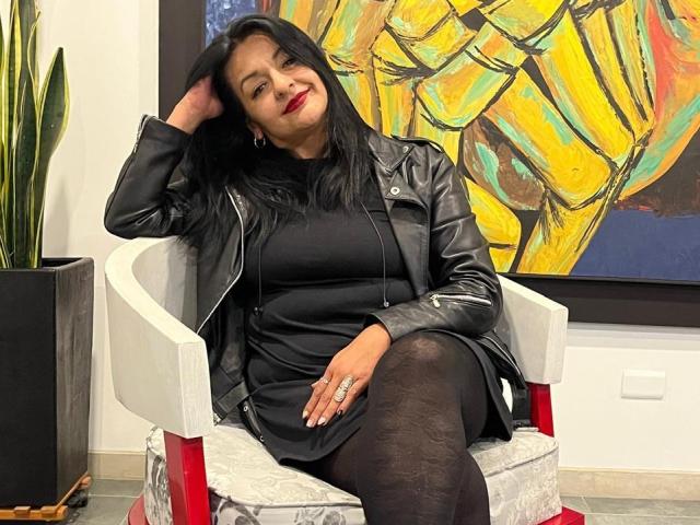 LiaDesire with black hair, red lipstick, black leather jacket, black dress, and black patterned tights, sitting on white chair, hand in hair, smiling, ring visible on finger.