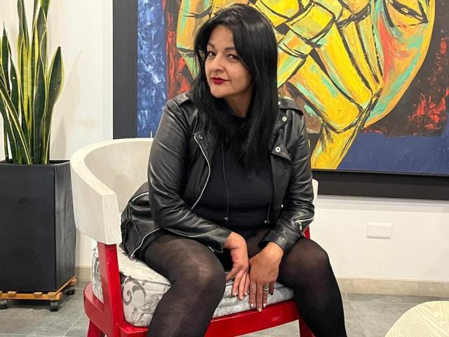 LiaDesire sits on a red chair wearing a black leather jacket, black top, and black tights. She has black hair, red lipstick, and is looking to the side.