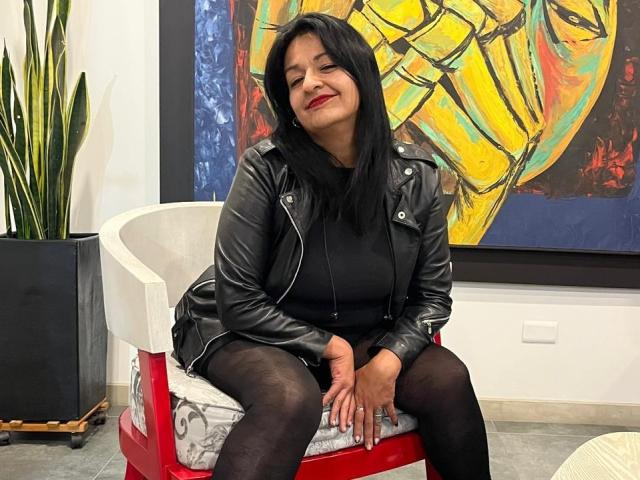 LiaDesire, black-haired woman, smiling, wearing black leather jacket, black top, black shorts, black tights, sitting on red chair, hands on lap, medium tits, red lipstick, indoor setting.