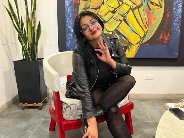 LiaDesire, with dark hair and glasses, sits on a red chair, wearing a black leather jacket and black pantyhose, touching her cheek, smiling. Her ass is covered.