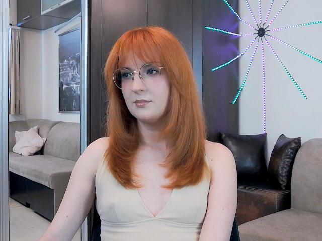 MoonBlossom with red hair, glasses, and pale skin, wearing a beige V-neck top, showing small tits, sitting in a modern room with a dark wall and neon star.