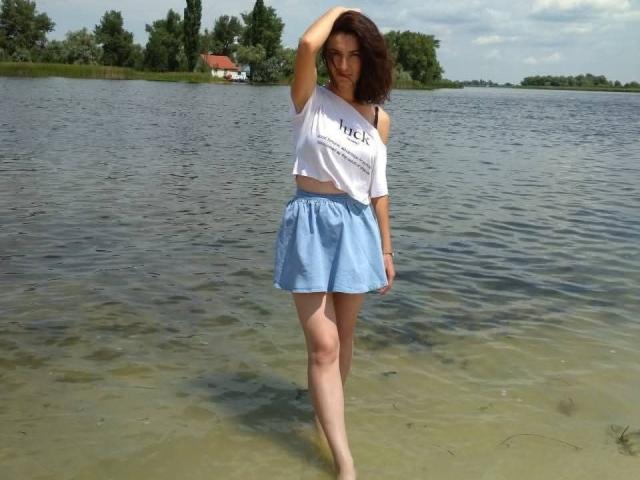 KatalinaQueen69 with brown hair, wearing a white crop top and blue skirt, standing in clear water, hand in hair, showing her ass, looking down.