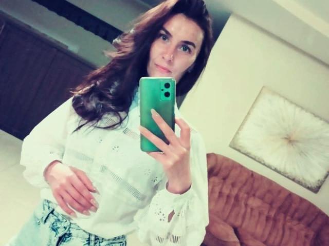 KatalinaQueen69 with wavy brown hair, wearing a white long-sleeve top and blue patterned pants, holding a green phone, taking a mirror selfie, showing a slight belly.