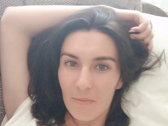 KatalinaQueen69 has pale skin, dark wavy hair, green eyes, and a relaxed, slightly smiling face with her right arm raised, lying on a white pillow.