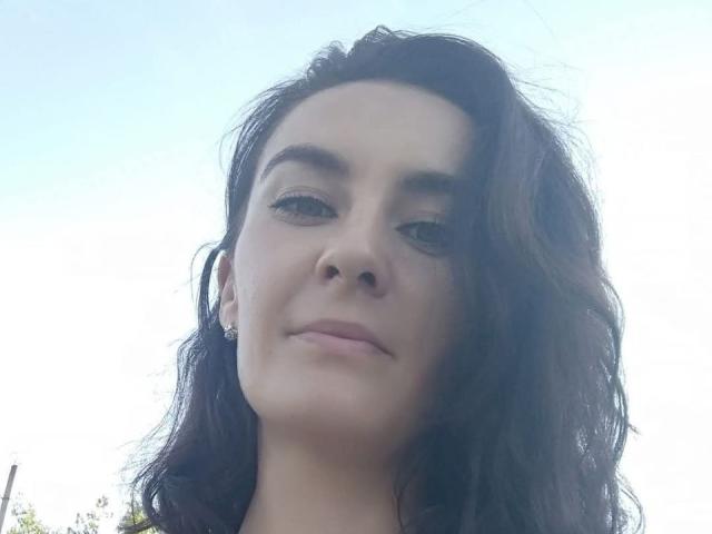 KatalinaQueen69 has wavy dark brown hair, light skin, and a slight smile with subtle makeup. She's wearing small earrings and looking down at the camera.