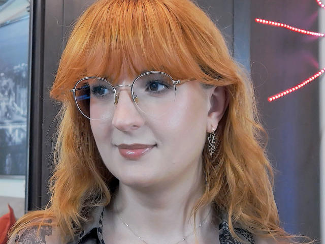 MoonBlossom has orange hair, blue eyes, and wears round glasses with metal frames. She's smiling with glossy pink lips, wearing a black patterned top and a silver earring.