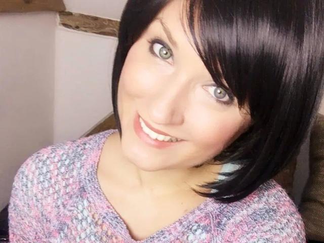 A MissDoertie-hot tem cabelo preto curto, olhos verdes e um sorriso adorável. Ela está vestindo um suéter rosa e cinza de tricô, que realça suas nádegas com um leve movimento..