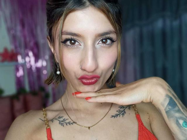 Kaiix with red lipstick, nose ring, and tattoos, pouting with hand on chin, wearing red top, has red nails, and brown hair, looking at camera with sultry eyes.