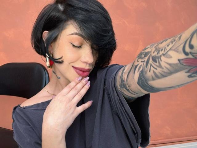 HannaBorbon with short black hair, tattoos on her right arm, dark shirt, purple nails, red lipstick, smiling, hand near face, black chair, brown-orange wall.