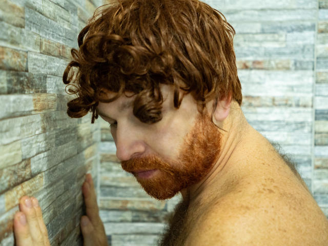 Michael Cosio, com cabelo ruivo encaracolado e barba, sardento, sem camisa, pressionado contra uma parede de pedra, olhando para baixo com uma expressão séria, a mão direita tocando a parede..