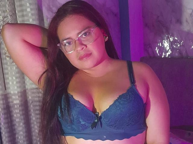 Chizuqui with glasses, dark long hair, blue lace bra, and a hand behind her head, showing her tits and a slightly pouty face in purple and pink lighting.
