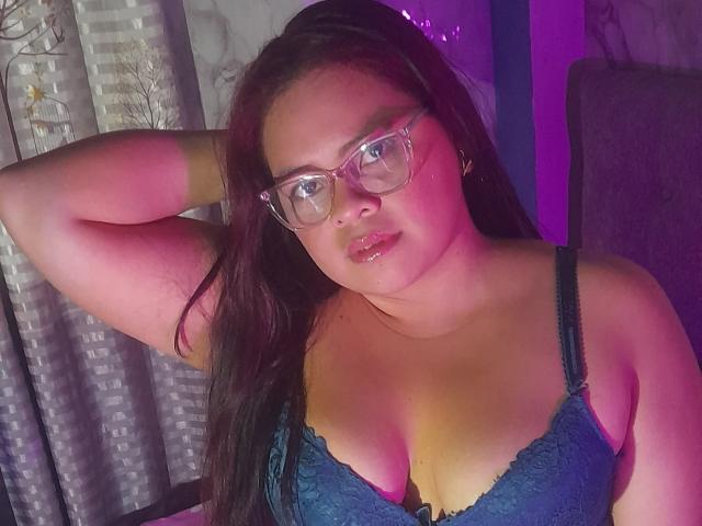 Chizuqui with glasses, dark hair, and shiny lips, wearing a blue lace bra, has her arm raised, showing her tits, in a pink and purple lit room.